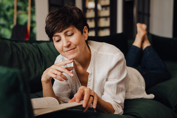 Smiling mature woman reading book while lying on couch