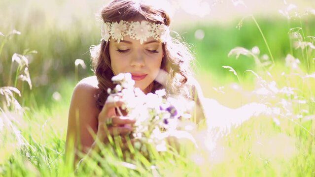 Beautiful hippie girl lying in green grass in a summer park holding some fresh wild flowers and wearing a vintage white lace headband with gentle sun flare