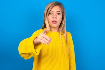 Shocked caucasian teen girl wearing yellow sweater over blue studio background points at you with stunned expression