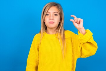 caucasian teen girl wearing yellow sweater over blue studio background purses lip and gestures with...