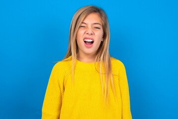 caucasian teen girl wearing yellow sweater over blue studio background yawns with opened mouth stands. Daily morning routine