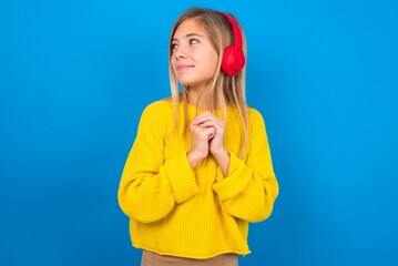 caucasian teen girl wearing yellow sweater over blue studio background wears stereo headphones listening to music concentrated and looking aside with interest.