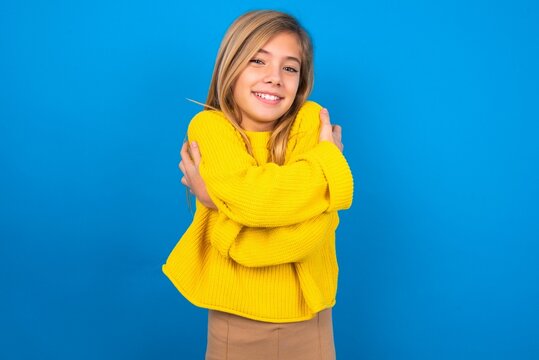 Charming Pleased Caucasian Teen Girl Wearing Yellow Sweater Over Blue Studio Background Embraces Own Body, Pleasantly Feels Comfortable Poses. Tenderness And Self Esteem Concept