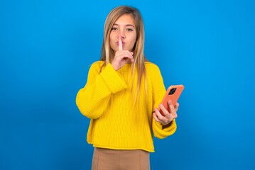 caucasian teen girl wearing yellow sweater over blue studio background holding modern gadget ask...