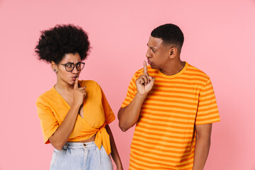 African couple showing silence gesture and looking at each other while standing isolated over pink background