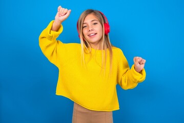 Carefree caucasian teen girl wearing yellow sweater over blue studio background with toothy smile raises arms dances carefree moves with rhythm of music listens music from playlist via headphones