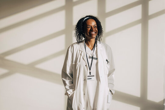 Smiling mature female physician with hands in pockets standing against wall with shadow