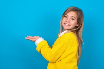 caucasian teen girl wearing yellow sweater over blue studio background pointing aside with hands open palms showing copy space, presenting advertisement smiling excited happy