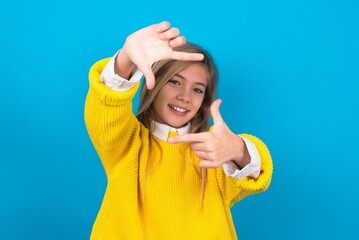 caucasian teen girl wearing yellow sweater over blue wall making finger frame with hands. Creativity and photography concept.