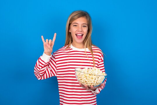 beautiful caucasian teen girl wearing striped T-shirt over blue wall eating popcorn doing a rock gesture and smiling to the camera. Ready to go to her favorite band concert.