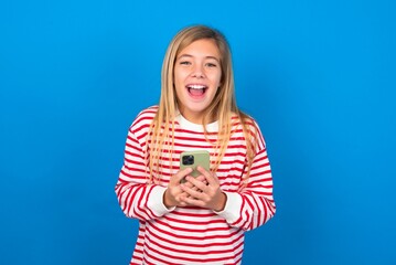 Excited beautiful caucasian teen girl wearing striped T-shirt over blue wall holding smartphone and looking amazed to the camera after receiving good news.