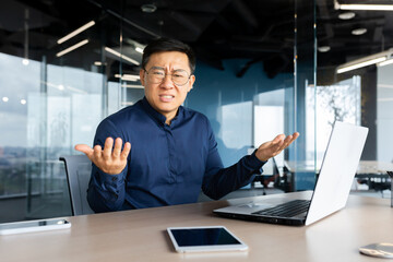 Portrait of frustrated and angry boss inside office, asian man looking at camera and shouting upset, man at workplace using laptop unhappy with work result.