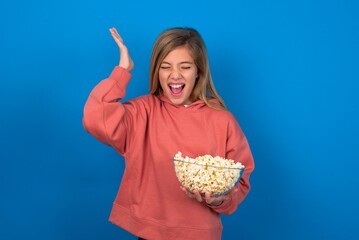 beautiful caucasian teen girl wearing pink sweater over blue wall eating popcorn goes crazy as head goes around feels stressed because of horrible situation