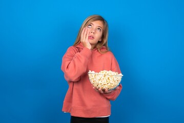 beautiful caucasian teen girl wearing pink sweater over blue wall eating popcorn keeps hands on cheeks has bored displeased expression. Stressed hopeless model