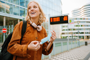 Cheerful woman putting on medical mask while standing at bus station