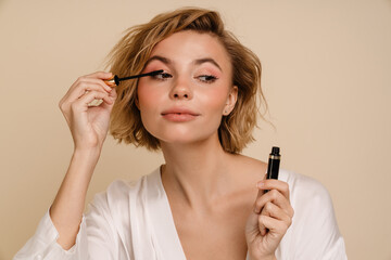 Young woman doing make up with mascara isolated over beige wall