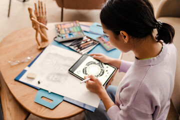 Back view of woman artist drawing on digital tablet while sitting in living room