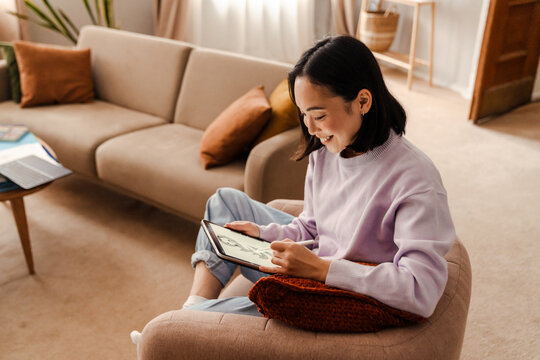 Joyful asian woman drawing on tablet with stylus while sitting at home