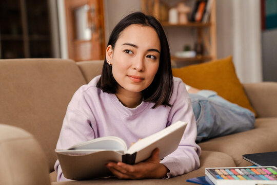 Thoughtful asian woman reading book while laying on couch