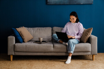 Cheerful asian woman working on laptop while sitting on couch