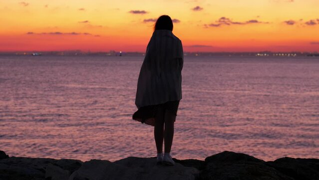 Woman in a light dress on waterfront, putting a blanket over her shoulders to keep warm. A strong breeze on a summer evening, Marmara Sea and colorful post-sunset sky visible in background
