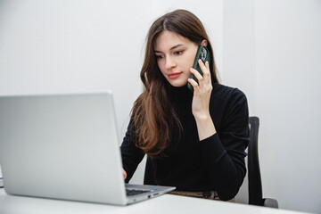 A young girl with long dark hair in black clothes sitting at a white table in the office and talks on the phone looking into a white laptop