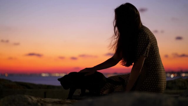 Woman stroking back of a stray cat on Istanbul's waterfront, silhouetted shot against sunset sky. Warm summer evening at city sea-front park at Maltepe.