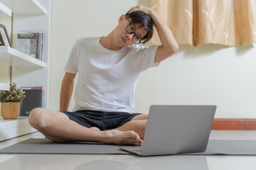 Heathy Asian man stretching neck, using laptop for learning yoga stretching for relax on online yoga class at home. Man stretching before workout at home. Home online workout yoga exercise concept - 569495854