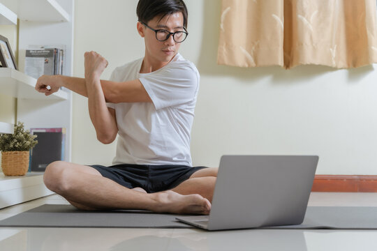 Heathy Asian man stretching shoulder, using laptop for learning yoga stretching for relax on online yoga class at home. Man stretching before workout at home. Home online workout yoga exercise concept