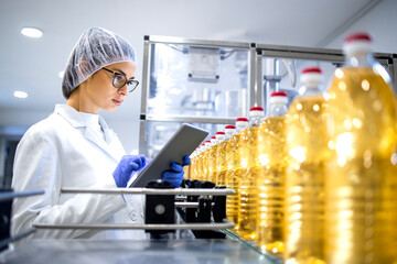 Food factory worker in sterile white uniform and hairnet using digital tablet while bottled refined oil is being produced on automated conveyor machine.