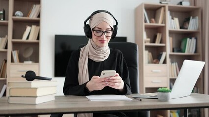Beautiful young muslim woman in headphones listening music and using phone, sitting at the desk at modern apartment. Pretty girl taking a break while listen music after hard work