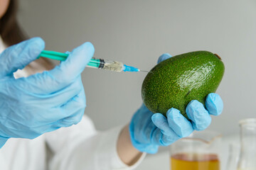 Young woman wearing scientist uniform injecting on avocado during experiment with vegetables in...
