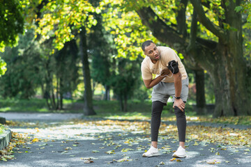Fototapeta premium Sick man has severe heart pain after jogging, african american man holding hand to chest fast heart rate, tired sportsman on sunny day in park, using headphones and phone to listen to music.