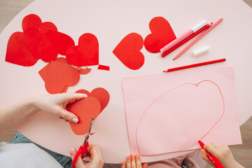 little girl and mother make Valentine's day cards using colored paper, scissors and pencil while...