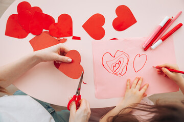 little girl and mother make Valentine's day cards using colored paper, scissors and pencil while sitting at a table