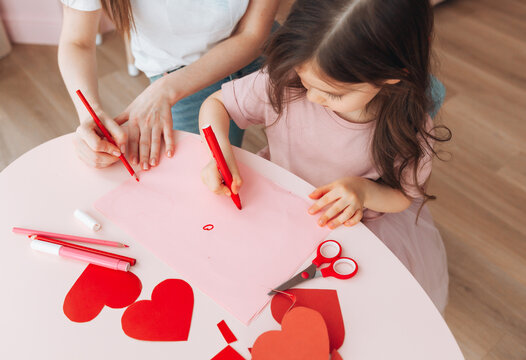 little girl and mother make Valentine's day cards using colored paper, scissors and pencil while sitting at a table