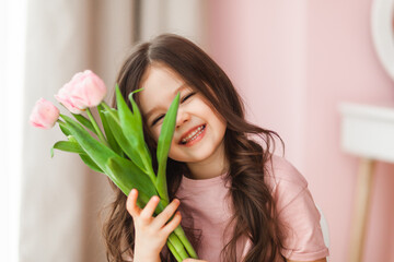 Naklejka premium Portrait of a little girl with long dark hair close-up. The baby hugs a bouquet of fresh, delicate pink tulips. A gift for the holiday, spring time.
