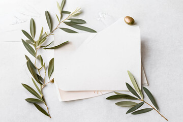 feminine minimalist styled wedding stationery mockup with a stack of blank invitation cards and a fresh olive twig on a white soft linen background, flat lay / top view