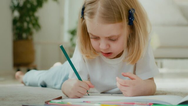 Cute creative funny preschool Caucasian little kid girl draw with colorful pencil lying on floor at home. Talented 5s child adopted daughter baby schoolgirl pupil drawing coloring picture painting
