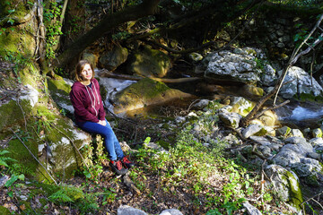 Obraz premium A smiling girl is resting on a rock near a picturesque forest stream during a hike