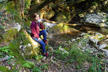 Naklejka na ściany i meble A girl resting in nature drinks a drink from a cup, sitting on a stone near a forest stream.