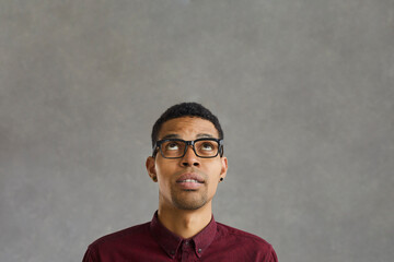 Dark-skinned man with doubtful facial expression looking up on gray concrete background. Man is...