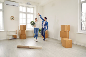 Young loving family couple having fun on moving day. Happy, cheerful man and woman dancing in a spacious and almost empty living room with several cardboard boxes in their new house or apartment