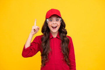 Worker teenager child wearing overalls red, cap and protect glasses. Studio shot portrait isolated on yellow background. Pointing and showing concept. Happy smiling emotions of teenager girl.
