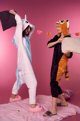Valentine's day concept. Studio portrait of two lesbians in kigurumi having a pillow fight at a pajama party. Cheerful girls are smiling and hitting each other with pillows on a pink background.