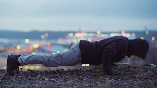 Exercising outdoors. Man doing push-ups on top of a hill. City lights in the background