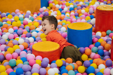 Fototapeta na wymiar A happy little boy has fun jumping into a dry pool with colorful balls. The kid boy is playing and having a good time in the game room with colorful balls.