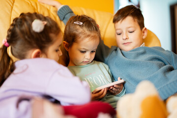 Сhildren sit on a leather sofa in the living room and watch cartoons on phone.