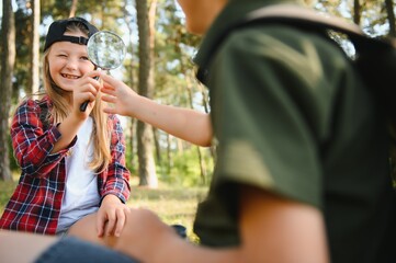 Group of curious happy school kids in casual clothes with backpacks exploring nature and forest together on sunny autumn day, girl holding magnifying glass and looking at fir cone in boy hands.