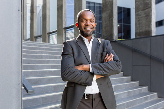 Portrait of a successful African American businessman. Standing in a suit outside the office center. He looks confidently into the camera, crosses his arms, smiles.